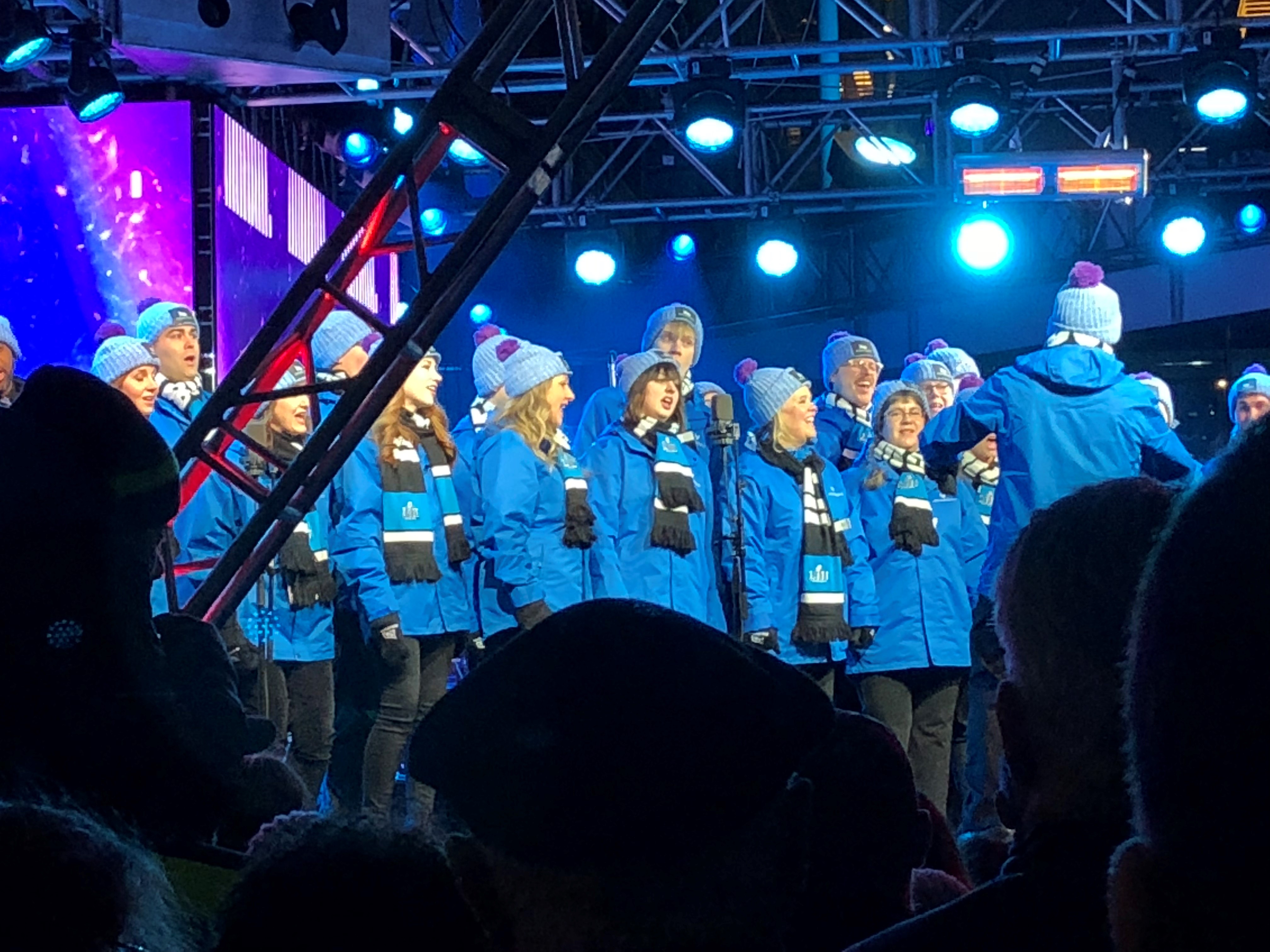 VocalEssence Singers and Philip Brunelle, wearing blue coats, blue and black scarves, and blue hats with purple pompoms perform for Super Bowl Live. Photo Credit Deb Vander Kooi