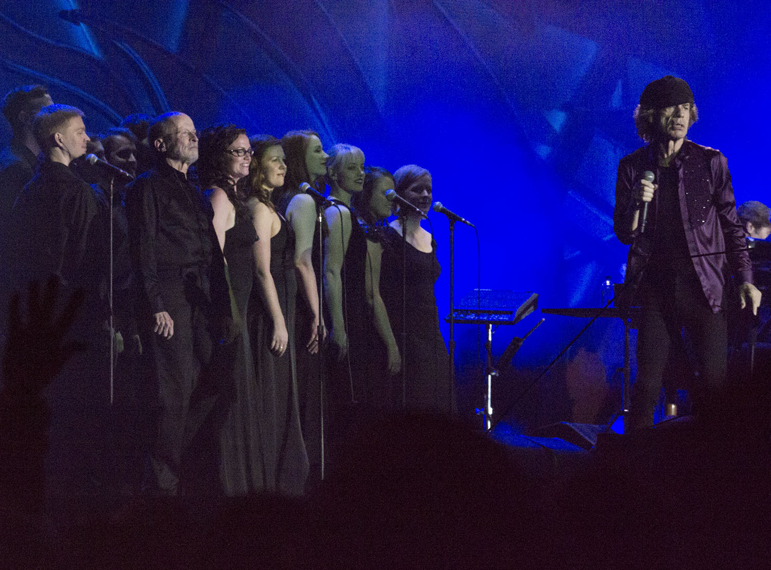 VocalEssence Singers wearing black stand and sing behind The Rolling Stones lead singer Mick Jagger, wearing a hat, a purple button down shirt, and black pants. Photo Credit Mike Wurm