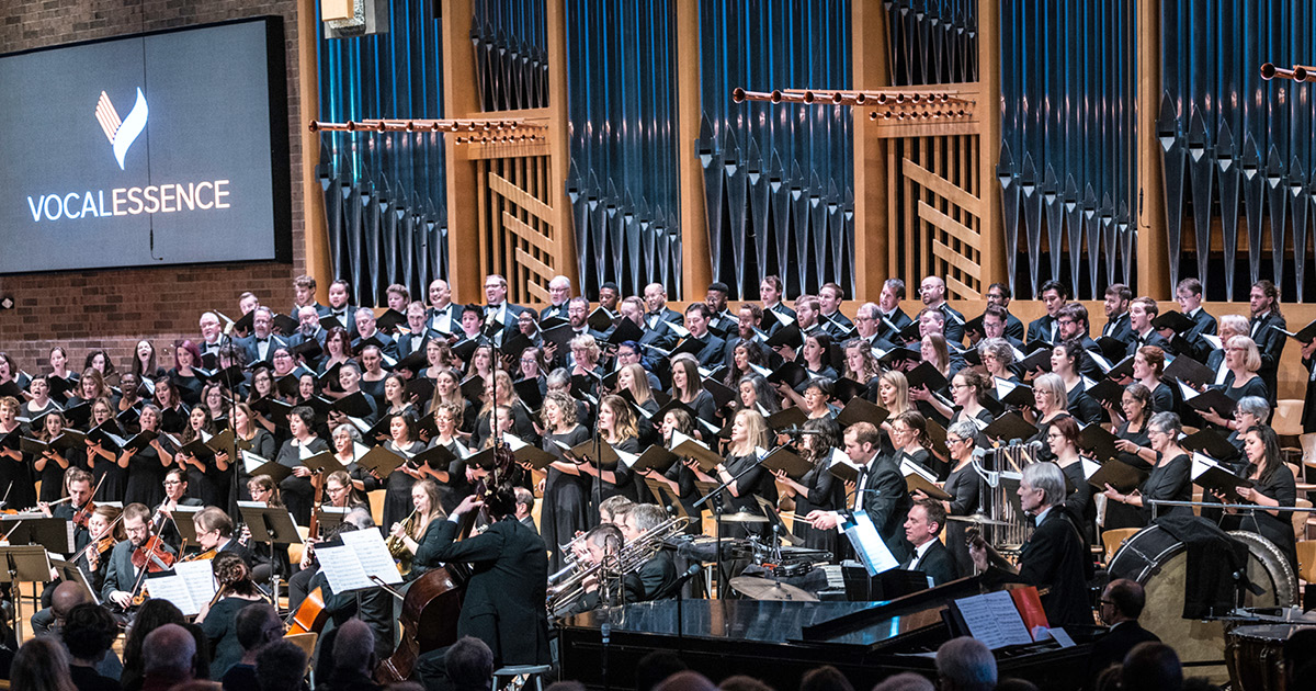 Members of VocalEssence holding black folders and singing with orchestra musicians in front of organ pipes with screen featuring VocalEssence logo in the upper left corner