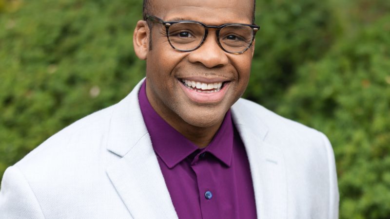 Person with glasses wearing a plum button down top, a light colored suit coat, and dark blue pocket square looking at the camera and smiling.