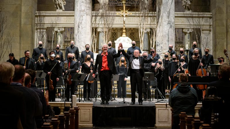 Man in black suit with red shirt and man in black tuxedo hold hands above their heads in front of orchestra musicans and singers. Photo Credit: Bruce Silcox