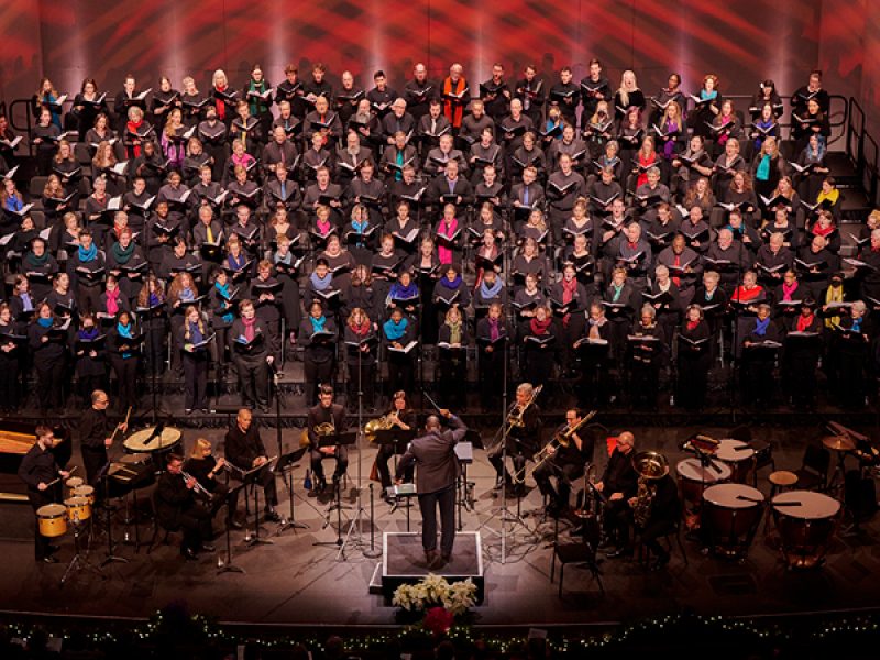 VocalEssence singers holding black folders performing at Northrop with brass and percussion players conducted by G. Phillip Shoultz, III. Photo Credit: Tony Nelson Photography