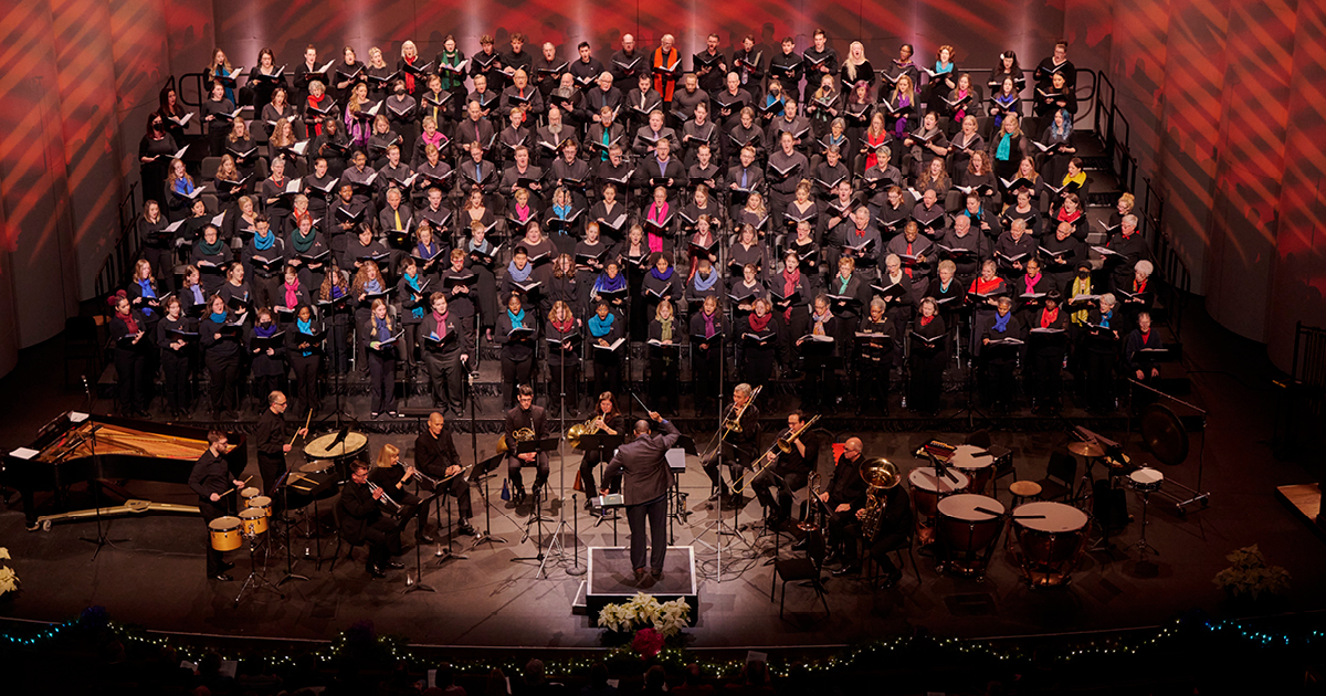 VocalEssence singers holding black folders performing at Northrop with brass and percussion players conducted by G. Phillip Shoultz, III. Photo Credit: Tony Nelson Photography