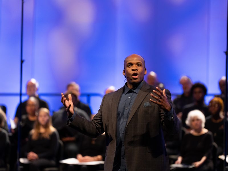 Person wearing a dark blue button down shirt and dark suit coat with hands reaching out and singing.