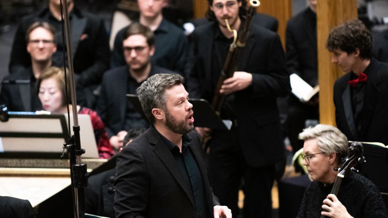 Person wearing black sings in front of orchestra players performing at a concert.