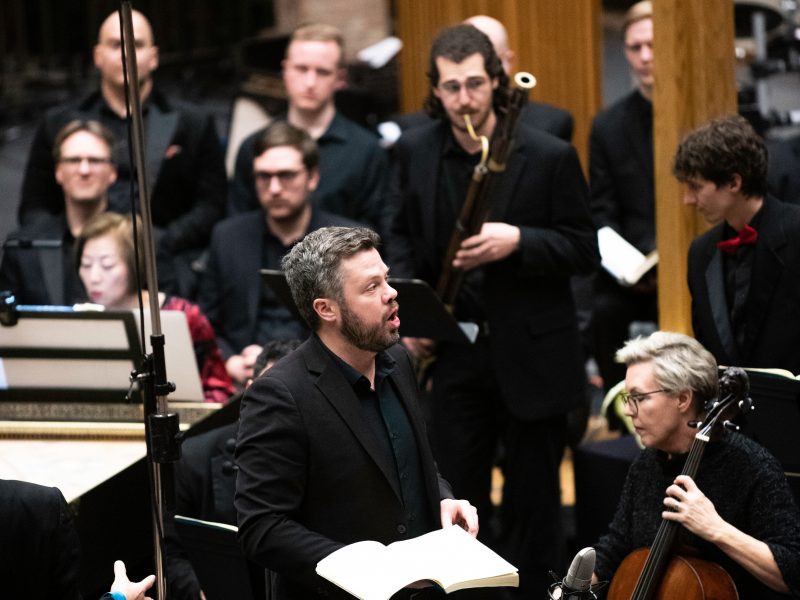 Person wearing black sings in front of orchestra players performing at a concert.