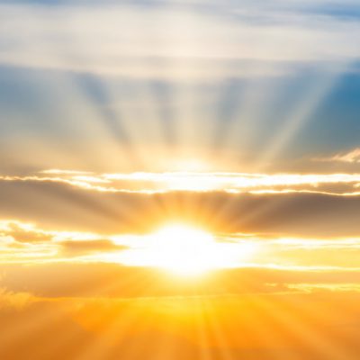 The sun and its rays shine beams of light through clouds breaking into blue sky above the sun and an orange hued sky below. Photo Credit: Adobe Stock Image