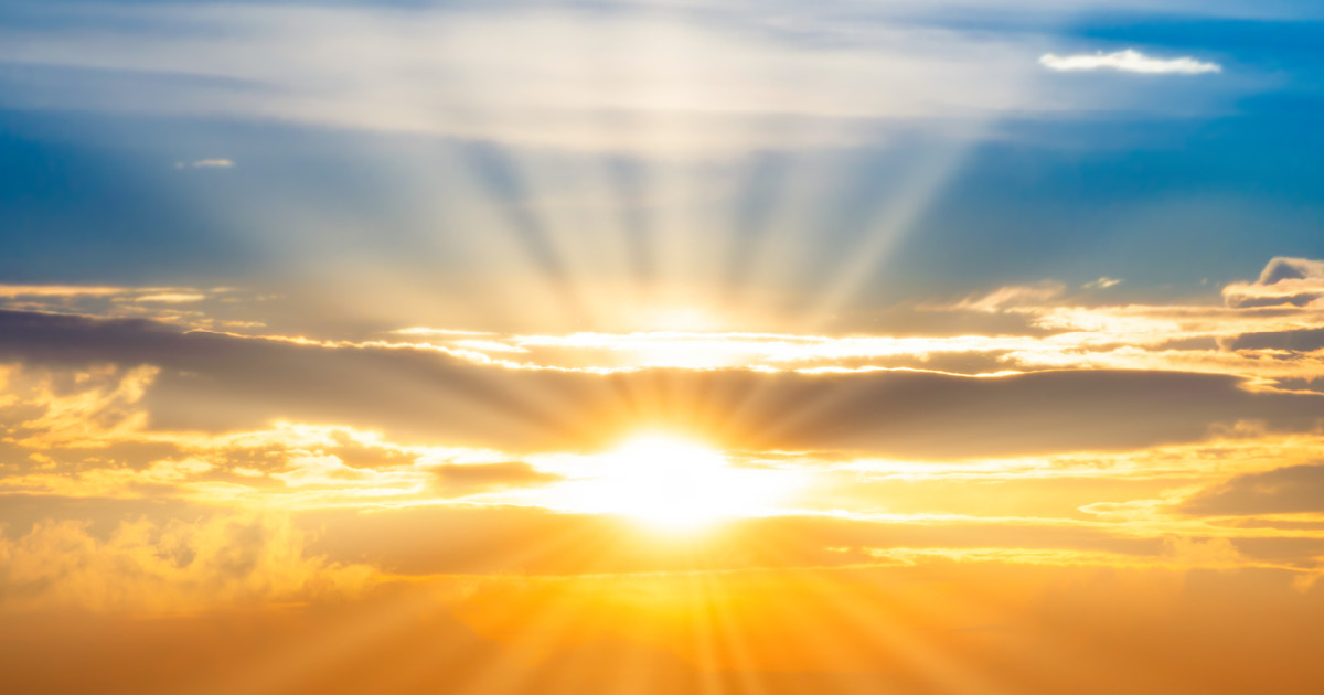 The sun and its rays shine beams of light through clouds breaking into blue sky above the sun and an orange hued sky below. Photo Credit: Adobe Stock Image