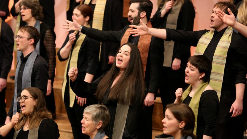 Singers wearing dark clothes with multicolored stoles sing with their hands reaching out.