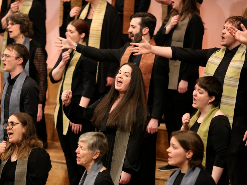 Singers wearing dark clothes with multicolored stoles sing with their hands reaching out.