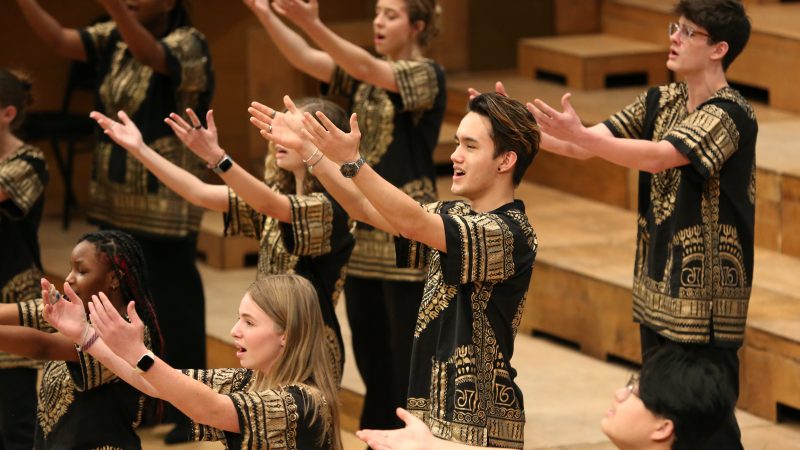Singers wearing dark clothes with a gold pattern sing with their hands reaching out.