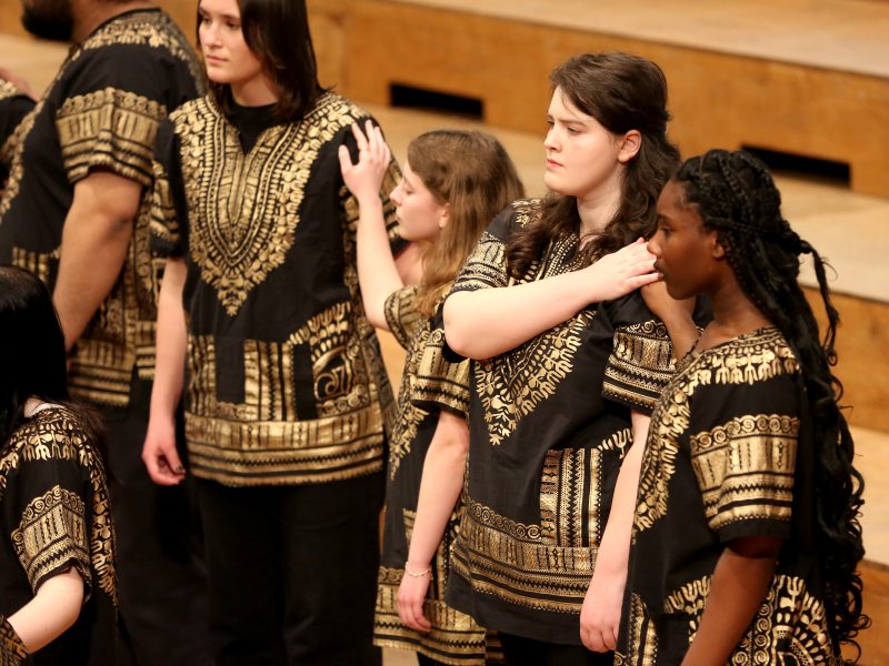 Singers wearing dark clothes with a gold pattern sing with their hands reaching out on the shoulders of the singer next to them.