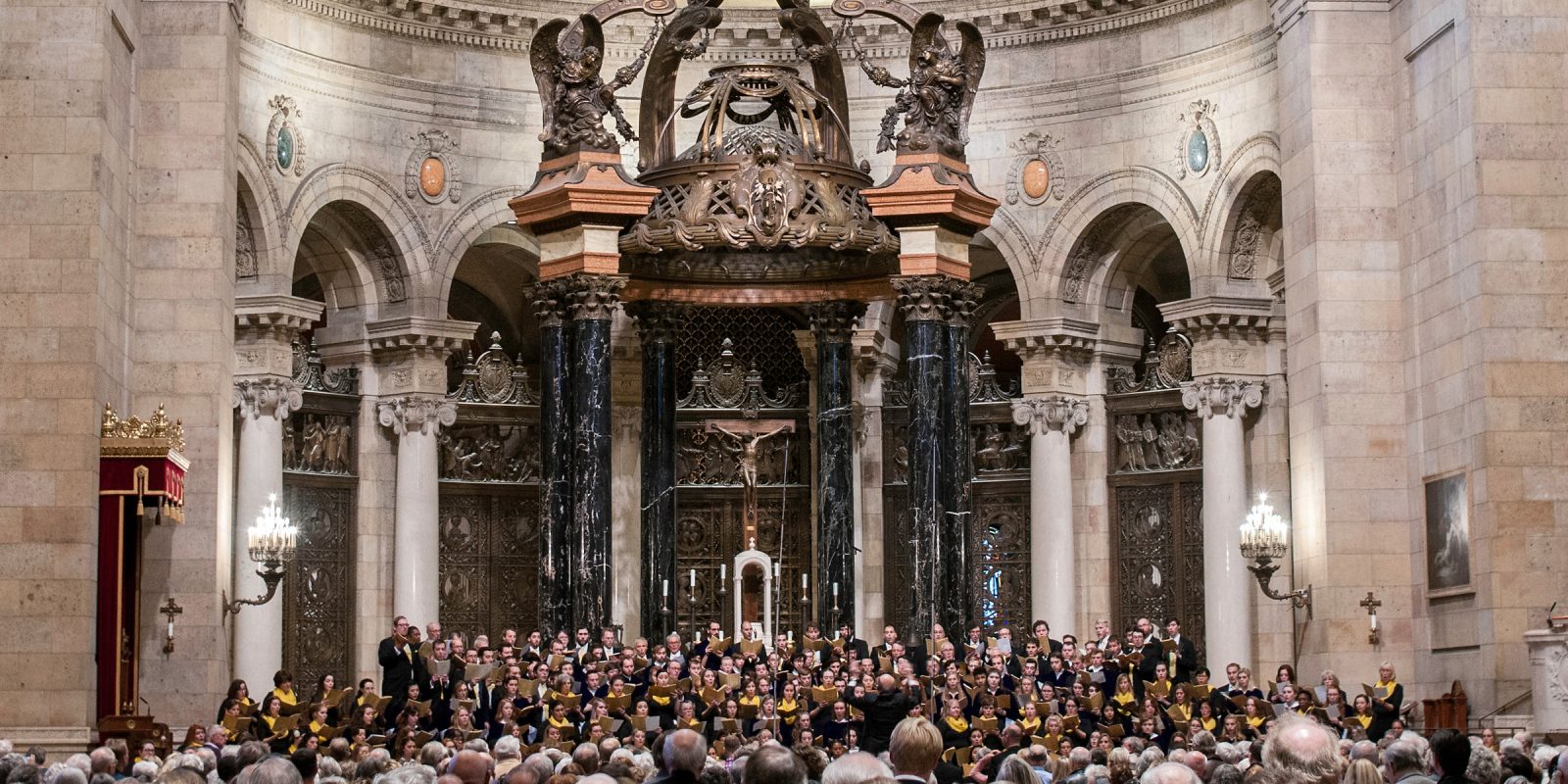 Many singers perform in front of an audience in the ornate sanctuary of the Cathedral of Saint Paul that features a baldachin with black and gold marble columns that has a bronze latticework canopy. Photo Credit: Bruce Silcox