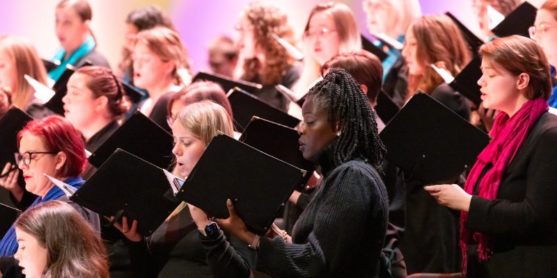 VocalEssence singers holding black folders and wearing black with colorful scarves perform at Northrop. Photo Credit: Anna Min