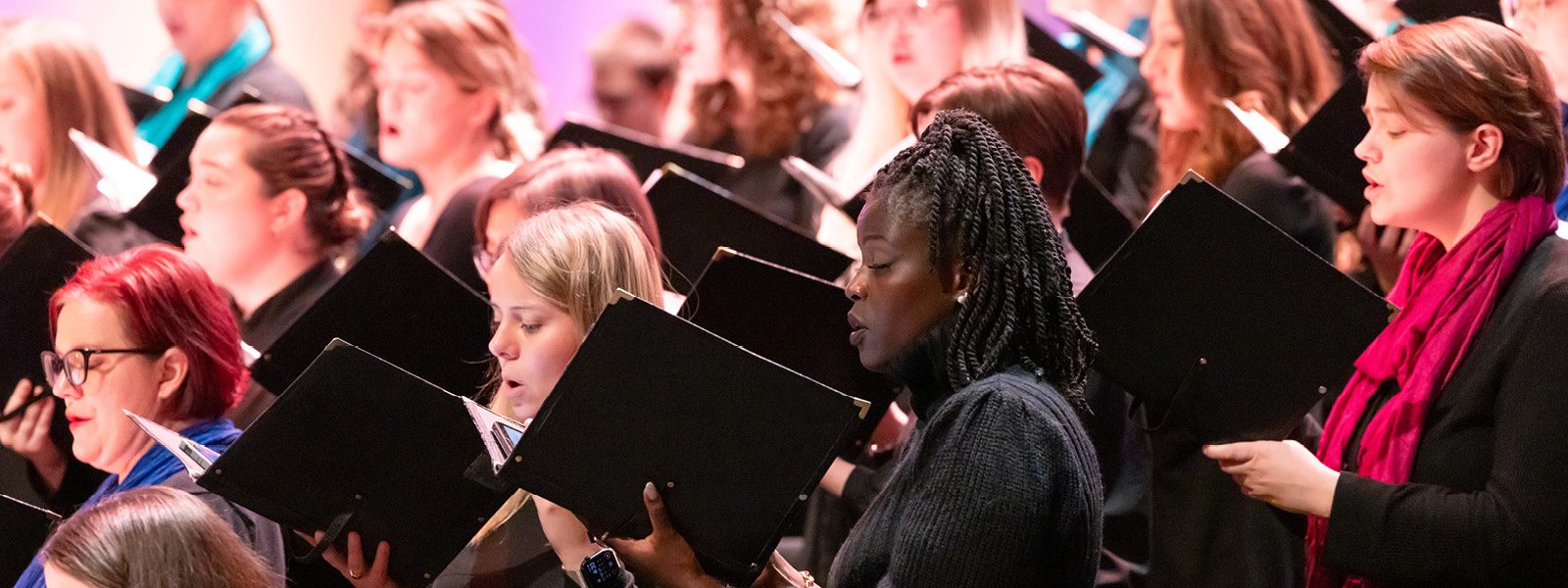 VocalEssence singers holding black folders and wearing black with colorful scarves perform at Northrop. Photo Credit: Anna Min