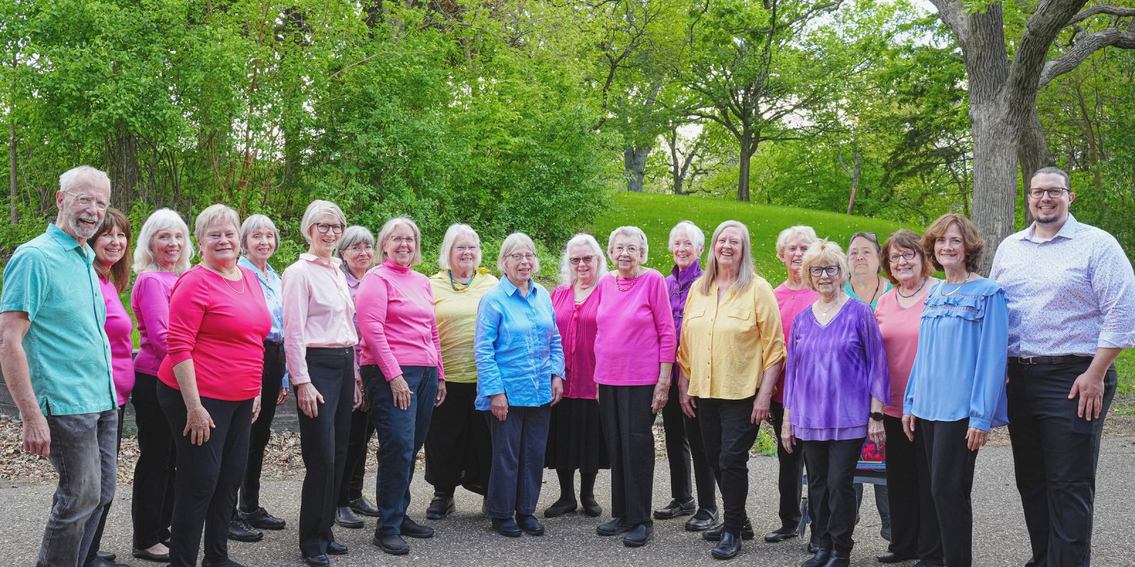 Adult Vintage Voices members wearing colorful shirts and black pants standing in a row and smiling standing in front of bright green trees and grass. Photo Credit: Novelli Jurado, Novart Media