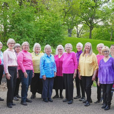 Adult Vintage Voices members wearing colorful shirts and black pants standing in a row and smiling standing in front of bright green trees and grass. Photo Credit: Novelli Jurado, Novart Media