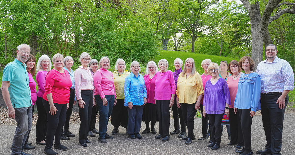 Adult Vintage Voices members wearing colorful shirts and black pants standing in a row and smiling standing in front of bright green trees and grass. Photo Credit: Novelli Jurado, Novart Media