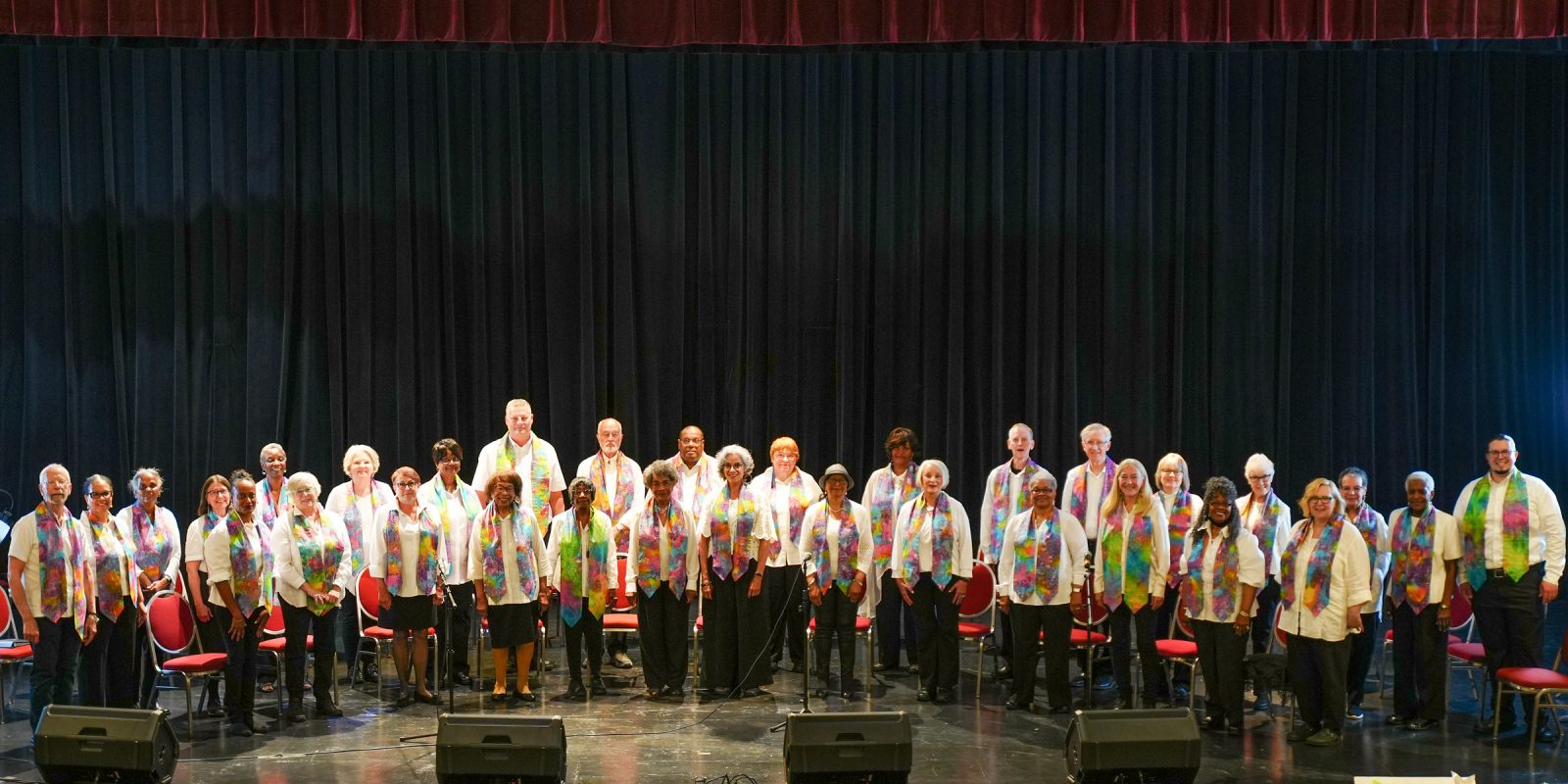 Adult Vintage Voices members wearing white shirts with colorful stoles and black pants standing in rows and smiling on an auditorium stage. Photo Credit: Novelli Jurado, Novart Media