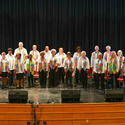 Adult Vintage Voices members wearing white shirts with colorful stoles and black pants standing in rows and smiling on an auditorium stage. Photo Credit: Novelli Jurado, Novart Media