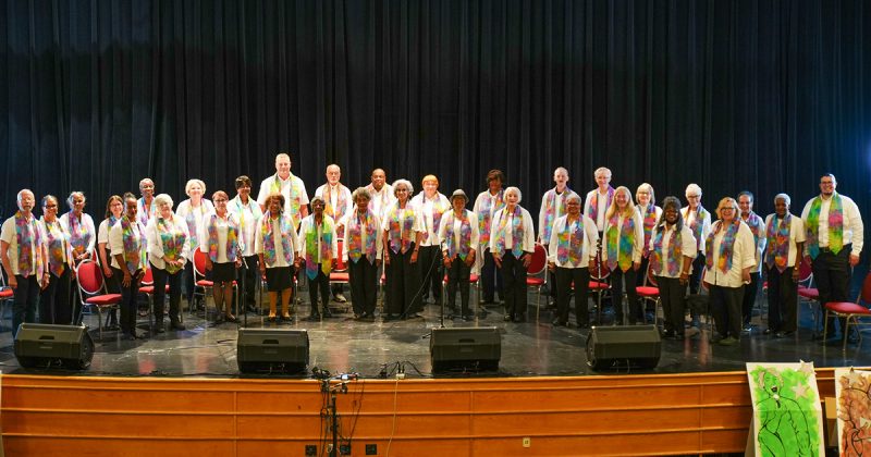 Adult Vintage Voices members wearing white shirts with colorful stoles and black pants standing in rows and smiling on an auditorium stage. Photo Credit: Novelli Jurado, Novart Media