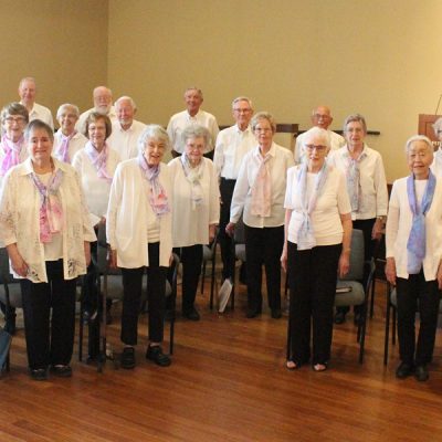 Adult Vintage Voices members wearing white shirts, black pants standing, and spring colored scarves in a row and smiling. Photo Credit: Trillium Woods Staff