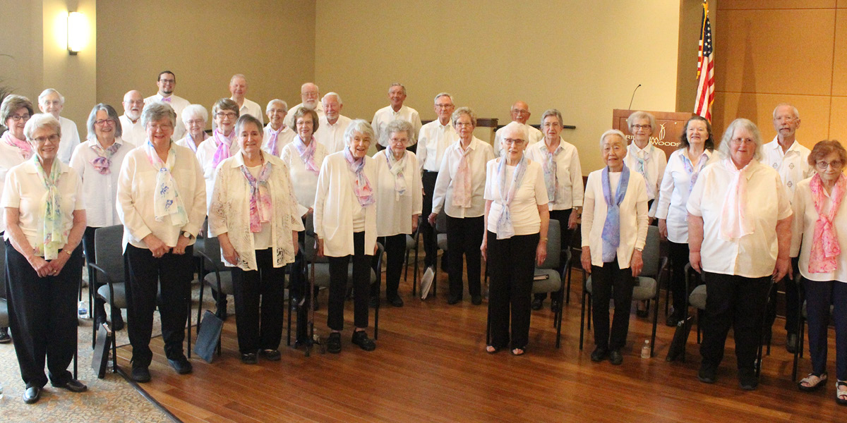 Adult Vintage Voices members wearing white shirts, black pants standing, and spring colored scarves in a row and smiling. Photo Credit: Trillium Woods Staff