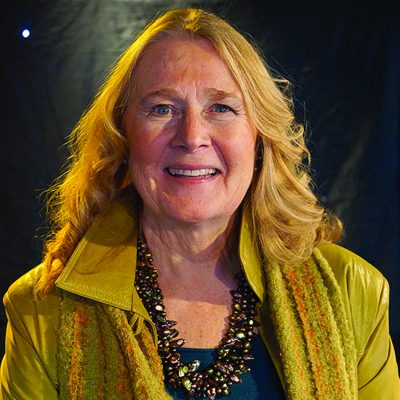 Headshot of Mary T who has shoulder-length, wavy blonde hair. She is smiling warmly and wearing a green jacket with a textured scarf in shades of green and yellow. 