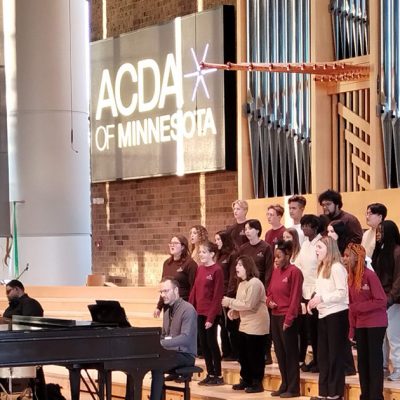 An image of singers, composed of diverse vocalists dressed in coordinated maroon and tan attire, performing under the direction of a conductor who stands at the front, guiding their performance. Musicians accompany the choir on instruments including a grand piano, guitar, and drums. Photo credit: Rhiannon Fiskradatz