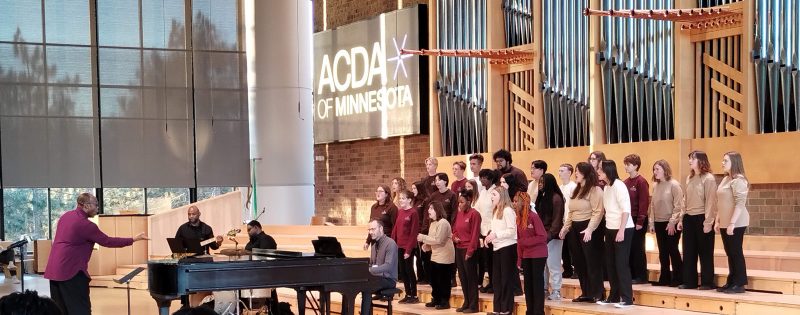 An image of singers, composed of diverse vocalists dressed in coordinated maroon and tan attire, performing under the direction of a conductor who stands at the front, guiding their performance. Musicians accompany the choir on instruments including a grand piano, guitar, and drums. Photo credit: Rhiannon Fiskradatz