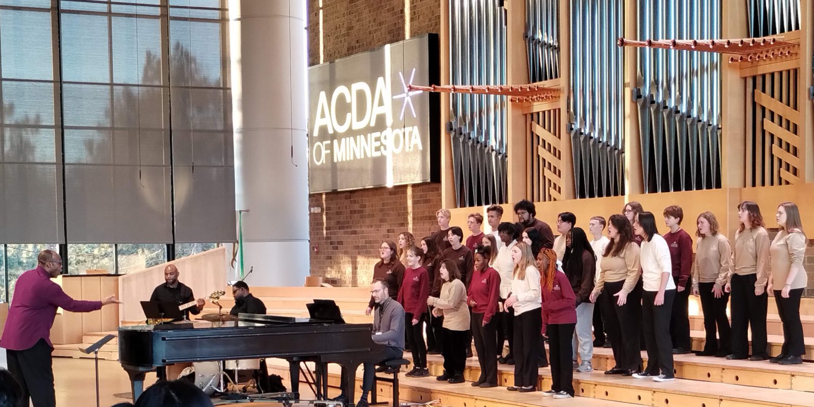 An image of singers, composed of diverse vocalists dressed in coordinated maroon and tan attire, performing under the direction of a conductor who stands at the front, guiding their performance. Musicians accompany the choir on instruments including a grand piano, guitar, and drums. Photo credit: Rhiannon Fiskradatz