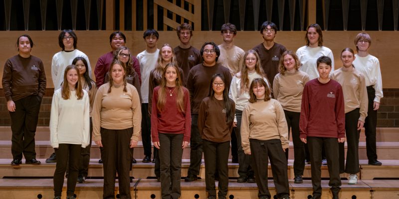 VocalEssence Singers Of This Age wearing different colored shirts and black pants standing on risers pose for a photo. Photo Credit: Ethan Kellum Johnson