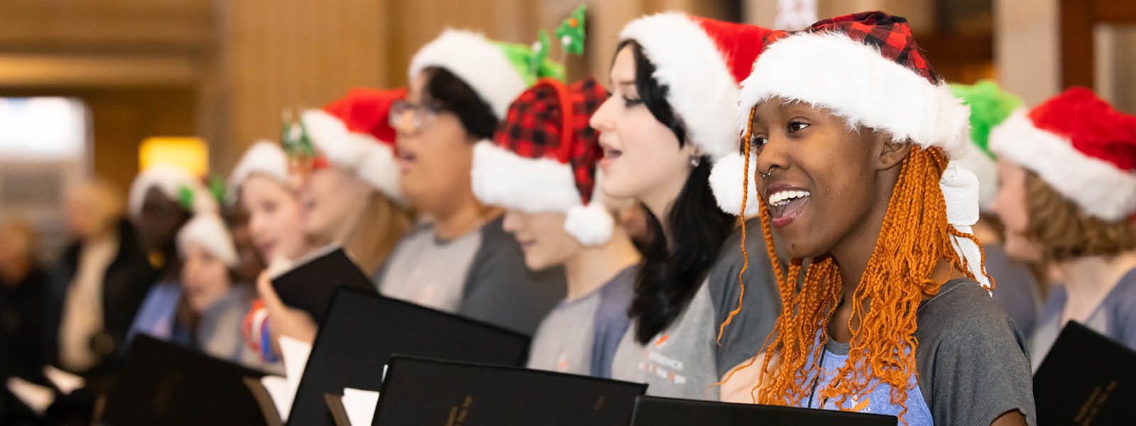 People wearing red and white Santa hats holding black folders and singing. Photo credit: Anna Min