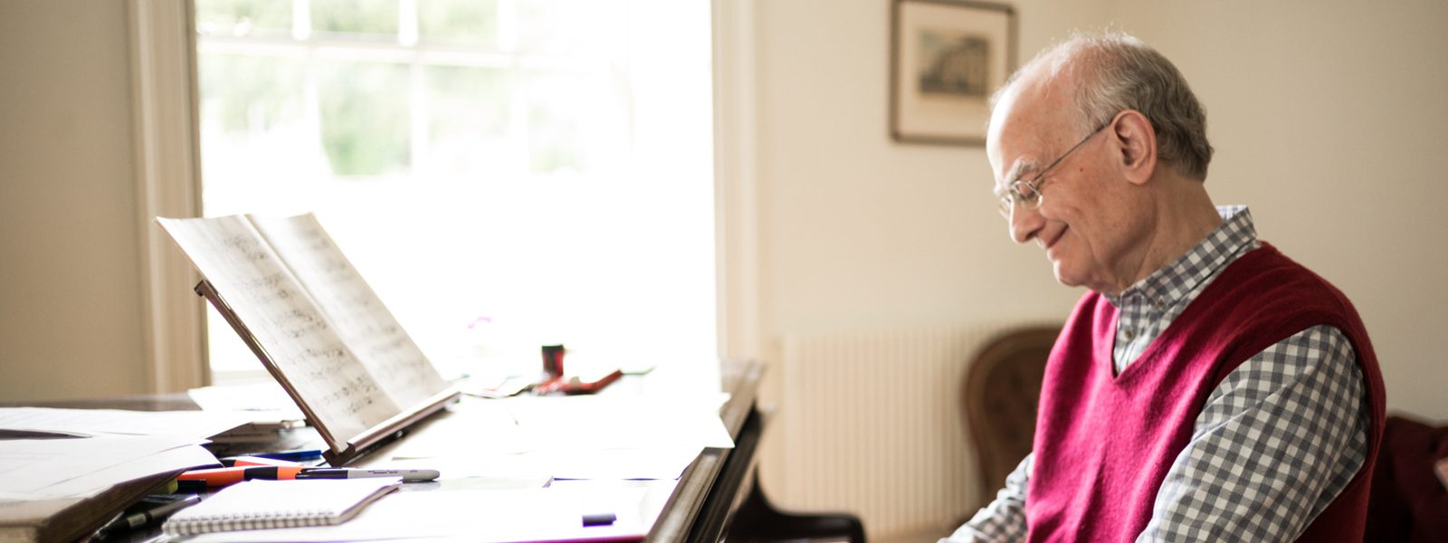John Rutter, wearing a red sweater vest over a checkered shirt, is smiling as he sits and plays the piano which has notebooks, pens, and sheet music. Photo credit: Unknown