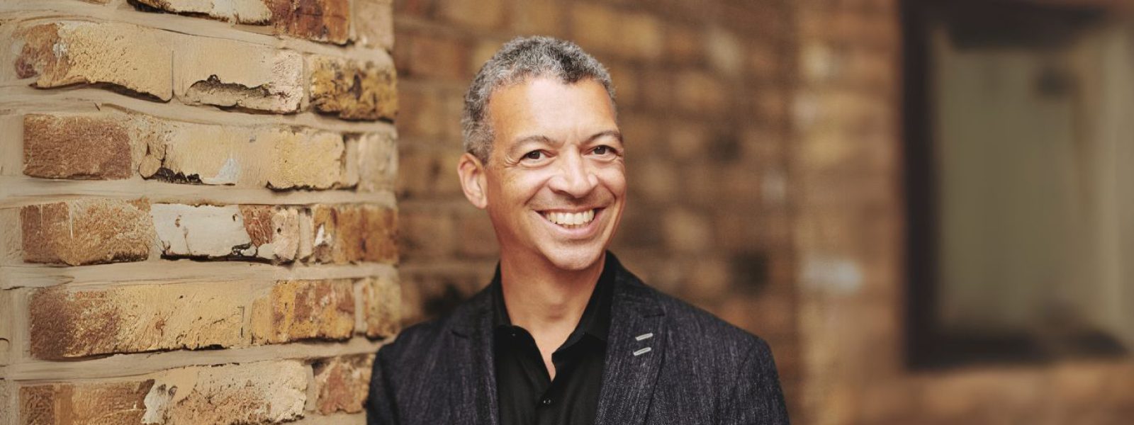 Roderick Williams, wearing a black button down shirt and a dark blazer with a full smile stands in front of a brick wall. Photo Credit: Theo Williams