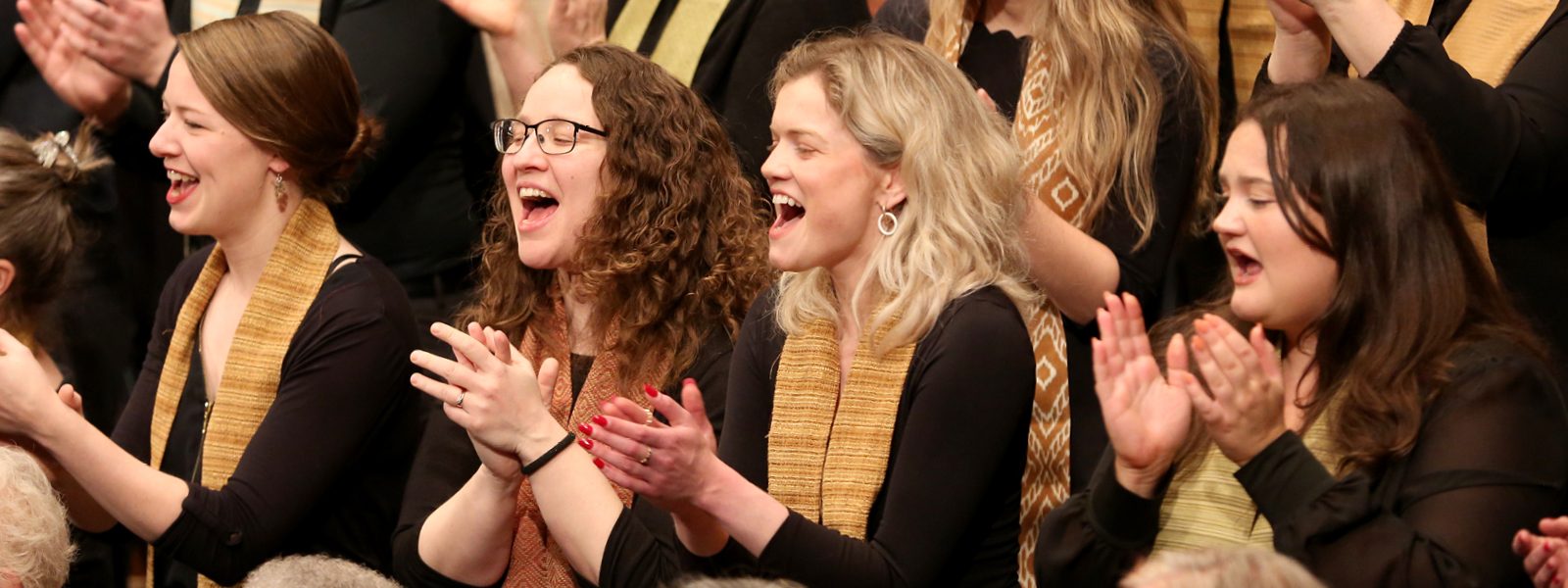 VocalEssence singers wearing black with multicolored stoles perform while clapping their hands with joyful expressions. Photo Credit: Kyndell Harkness
