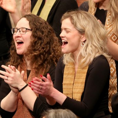 VocalEssence singers wearing black with multicolored stoles perform while clapping their hands with joyful expressions. Photo Credit: Kyndell Harkness