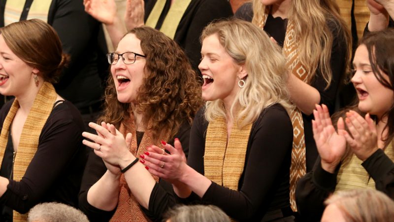 VocalEssence singers wearing black with multicolored stoles perform while clapping their hands with joyful expressions. Photo Credit: Kyndell Harkness