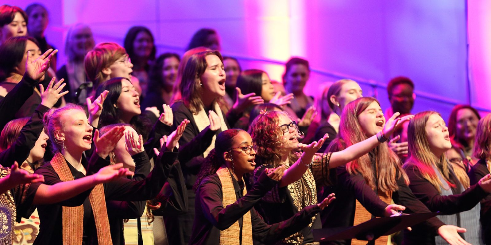 VocalEssence singers wearing black with multicolored stoles perform while holding their arms forward on stage for the annual WITNESS concert. Photo Credit: Kyndell Harkness
