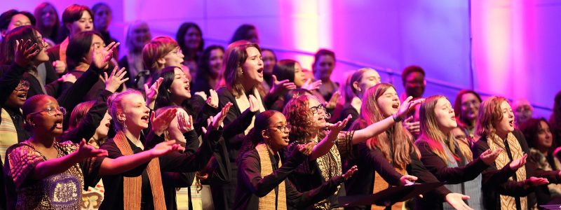 VocalEssence singers wearing black with multicolored stoles perform while holding their arms forward on stage for the annual WITNESS concert. Photo Credit: Kyndell Harkness