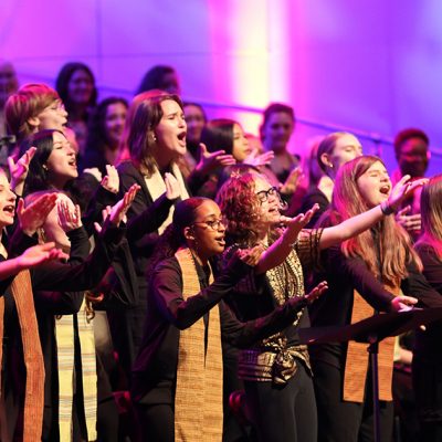 VocalEssence singers wearing black with multicolored stoles perform while holding their arms forward on stage for the annual WITNESS concert. Photo Credit: Kyndell Harkness