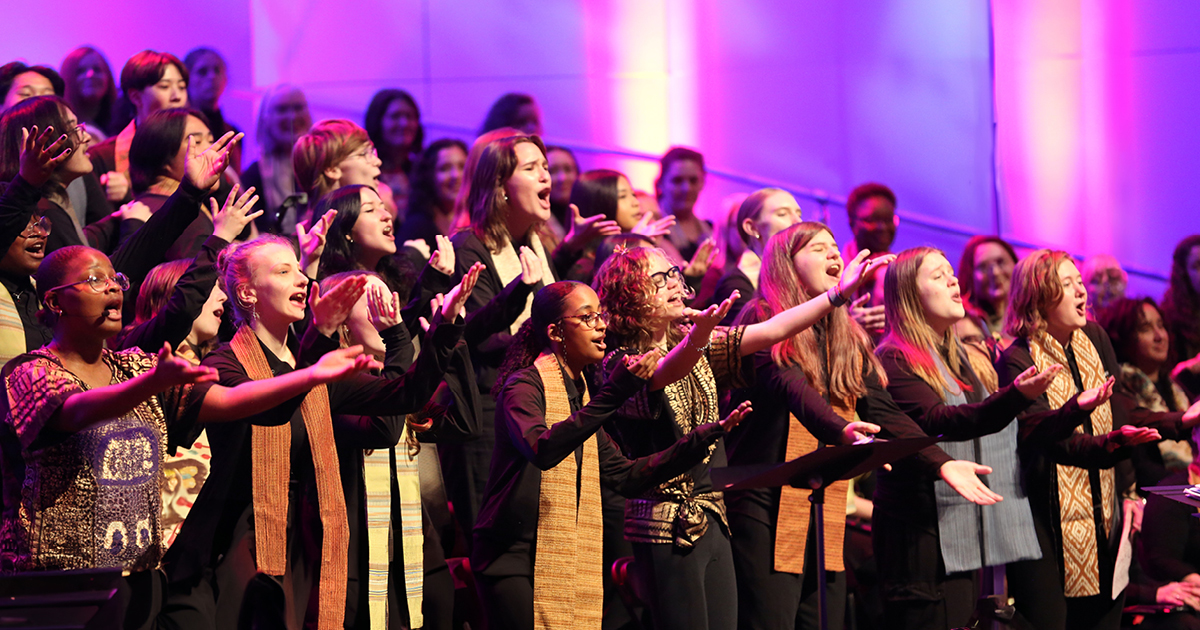 VocalEssence singers wearing black with multicolored stoles perform while holding their arms forward on stage for the annual WITNESS concert. Photo Credit: Kyndell Harkness