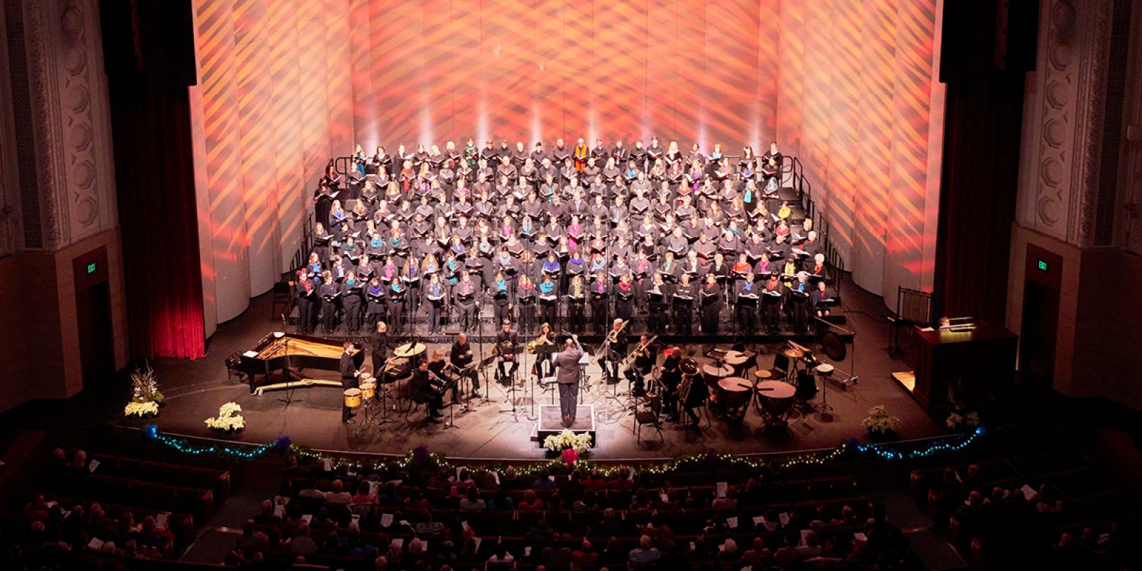 VocalEssence singers wearing black with colorful scarves holding black folders perform with brass and percussion on a poinsettia and twinkle light garland adorned stage at Northrop.