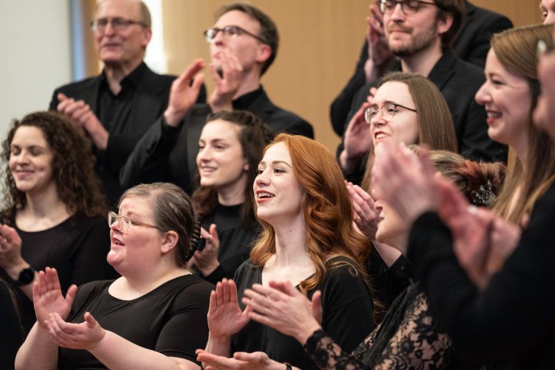 VocalEssence Ensemble Singers wearing black perform while clapping their hands with joyful expressions. Photo Credit: Bruce Silcox