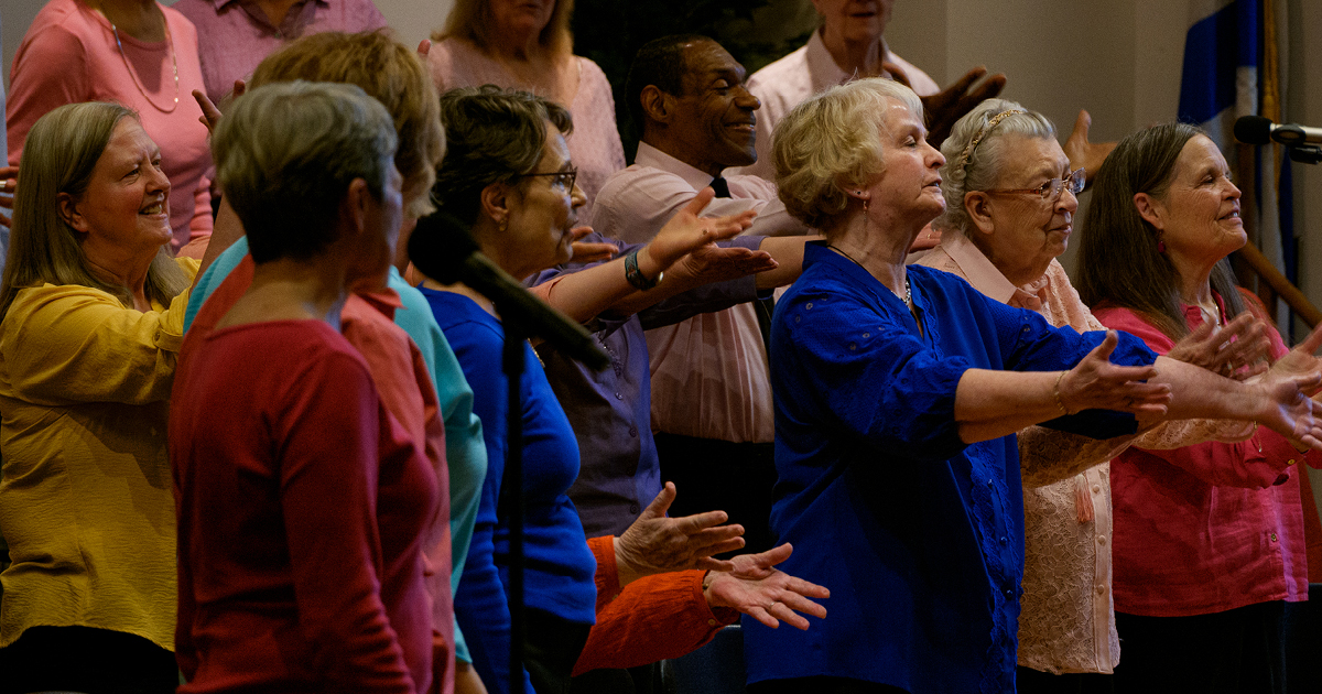 VocalEssence Vintage Voices singers wearing multicolored shirts sing joyfully while holding their arms forward. Photo Credit: Ethan Kellum Johnson