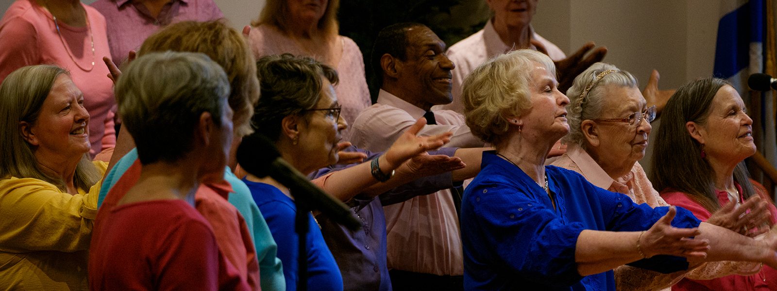 VocalEssence Vintage Voices singers wearing multicolored shirts sing joyfully while holding their arms forward. Photo Credit: Ethan Kellum Johnson