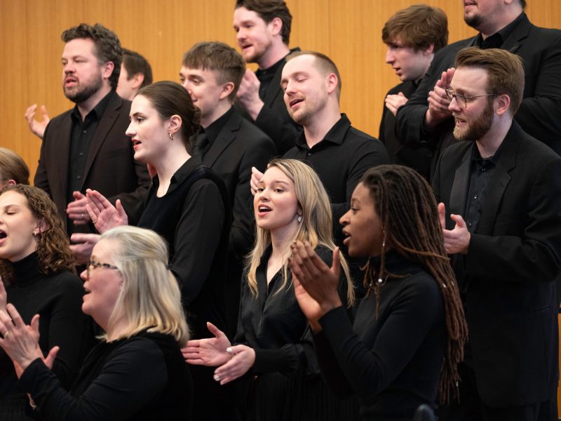 VocalEssence Ensemble Singers wearing black perform while clapping their hands with joyful expressions. Photo Credit: Bruce Silcox