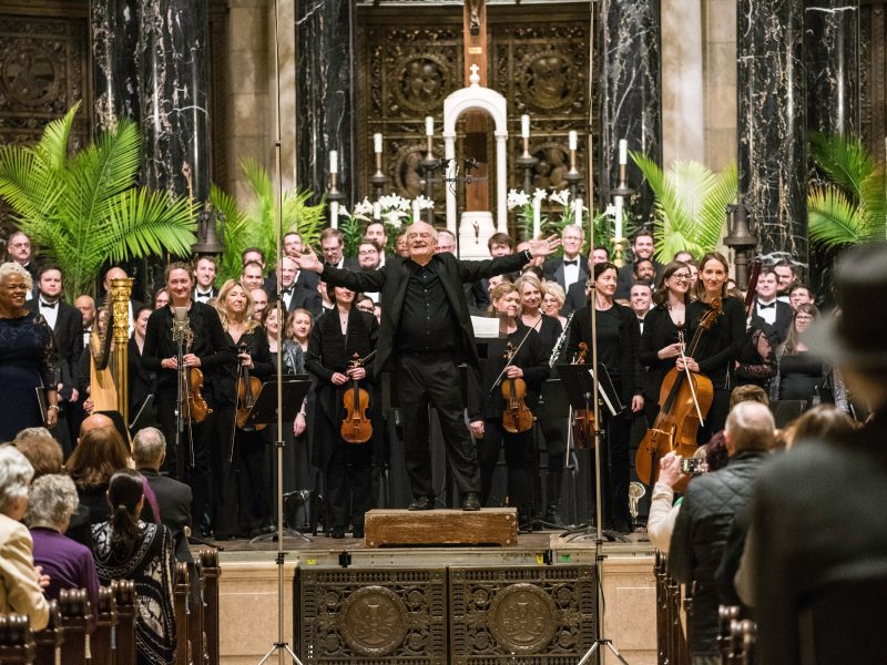 Person wearing black suit with arms outstretched stands in front of singers and orchestra in front of an audience in the ornate sanctuary of the Cathedral of Saint Paul. Photo Credit: Bruce Silcox