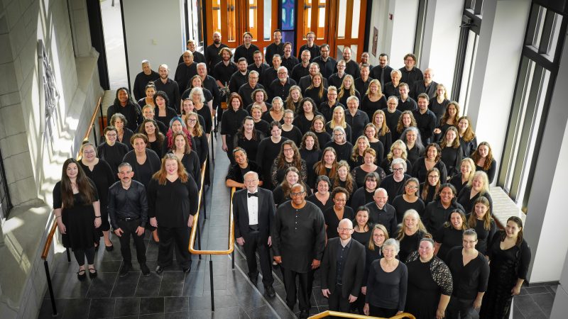 VocalEssence Chorus members wearing black pose for a photo in the lobby of a building with windows and stairs with gold railing. Photo Credit: Novart Media