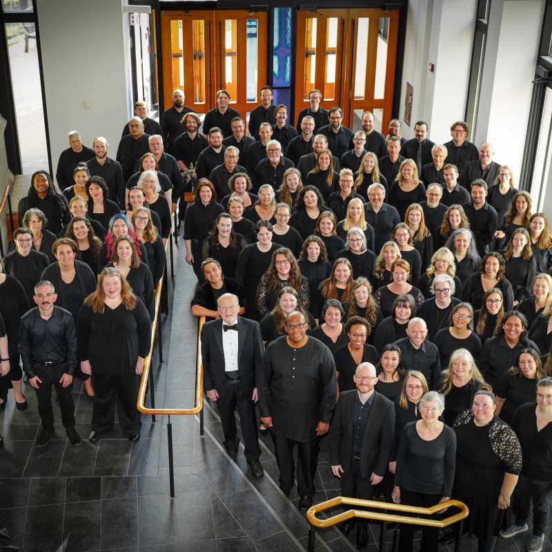 VocalEssence Chorus members wearing black pose for a photo in the lobby of a building with windows and stairs with gold railing. Photo Credit: Novart Media